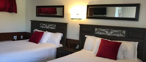 A hotel room with two beds, each with white bedding and red pillows, a nightstand with a clock, and a wall-mounted TV.