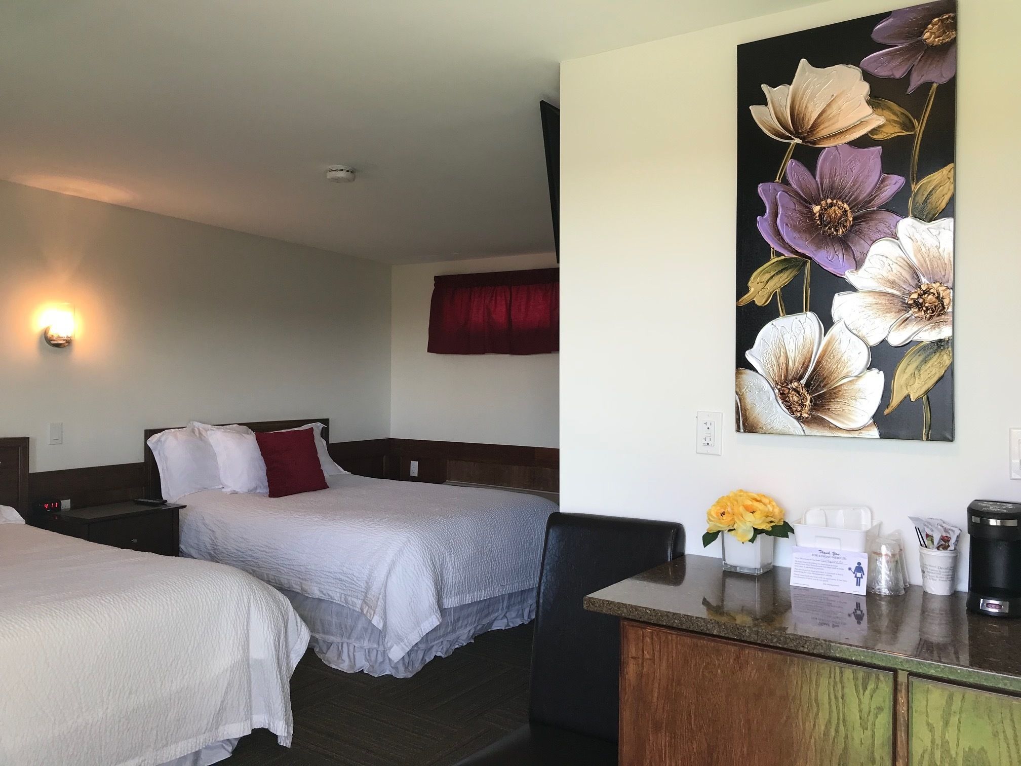 A hotel room with two beds, a desk, a coffee maker, and a floral painting on the wall.