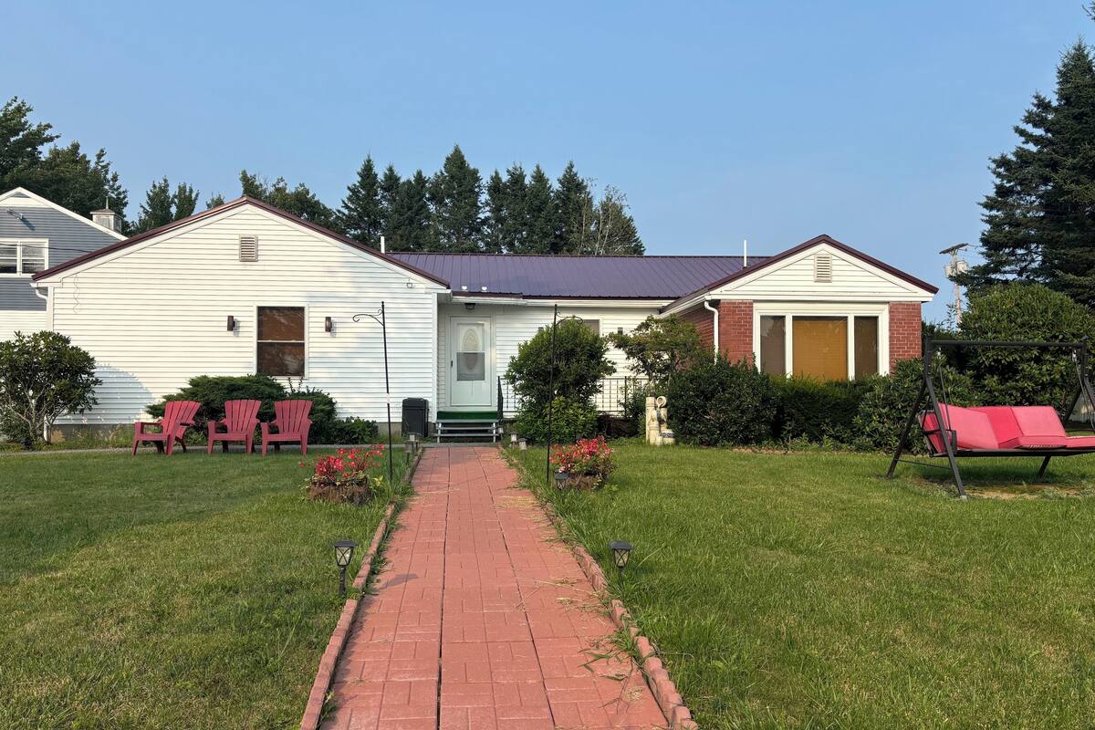 Une maison à deux étages, avec une partie en briques et une partie blanche, un chemin pavé menant à la porte d’entrée, et des chaises Adirondack rouges dans le jardin.