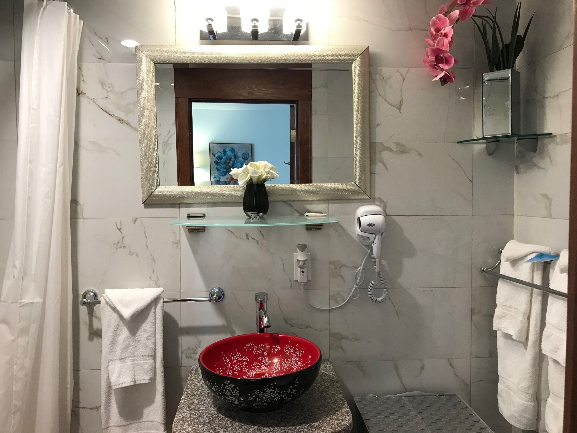 A bathroom with a marble-tiled wall, a large mirror, a red and black bowl sink, a hairdryer, and a vase with flowers.