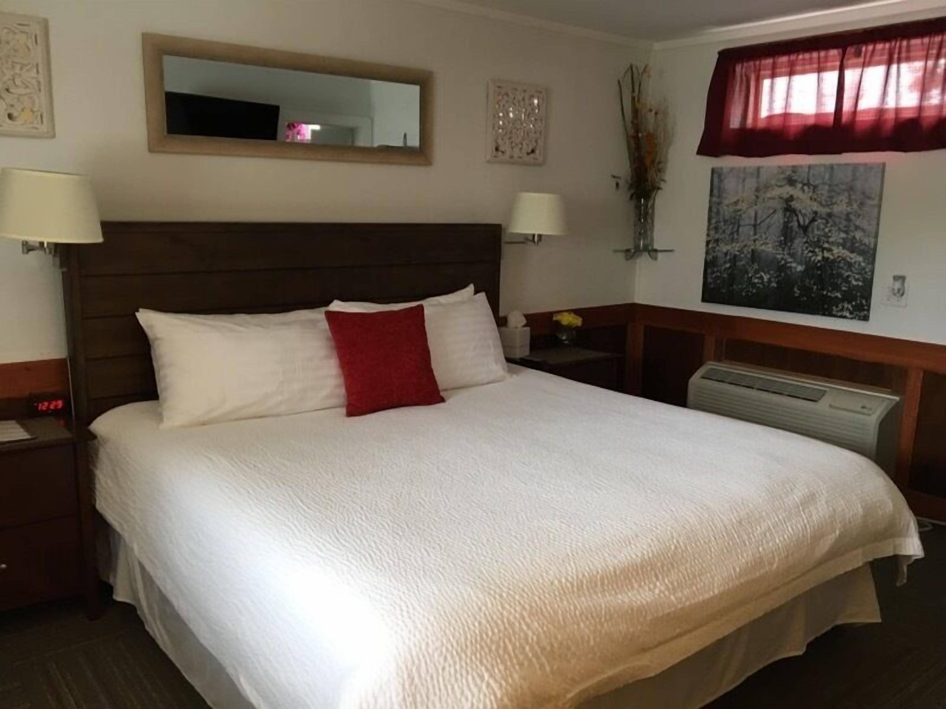A neatly made bed with white and red pillows, a wooden headboard, and a nightstand with a lamp.