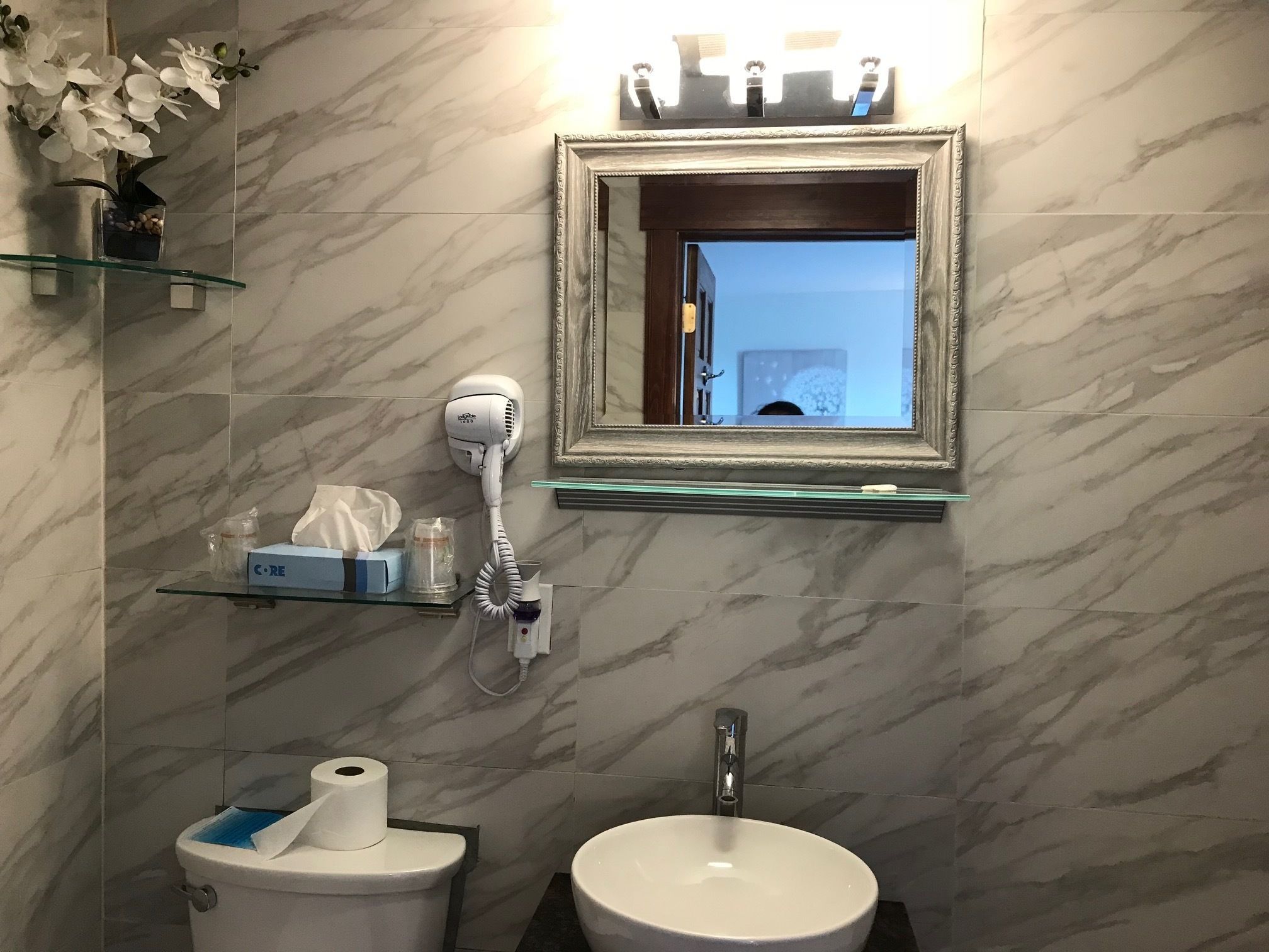 A bathroom with a marble wall, a white sink, a toilet, a hairdryer, and a framed mirror.