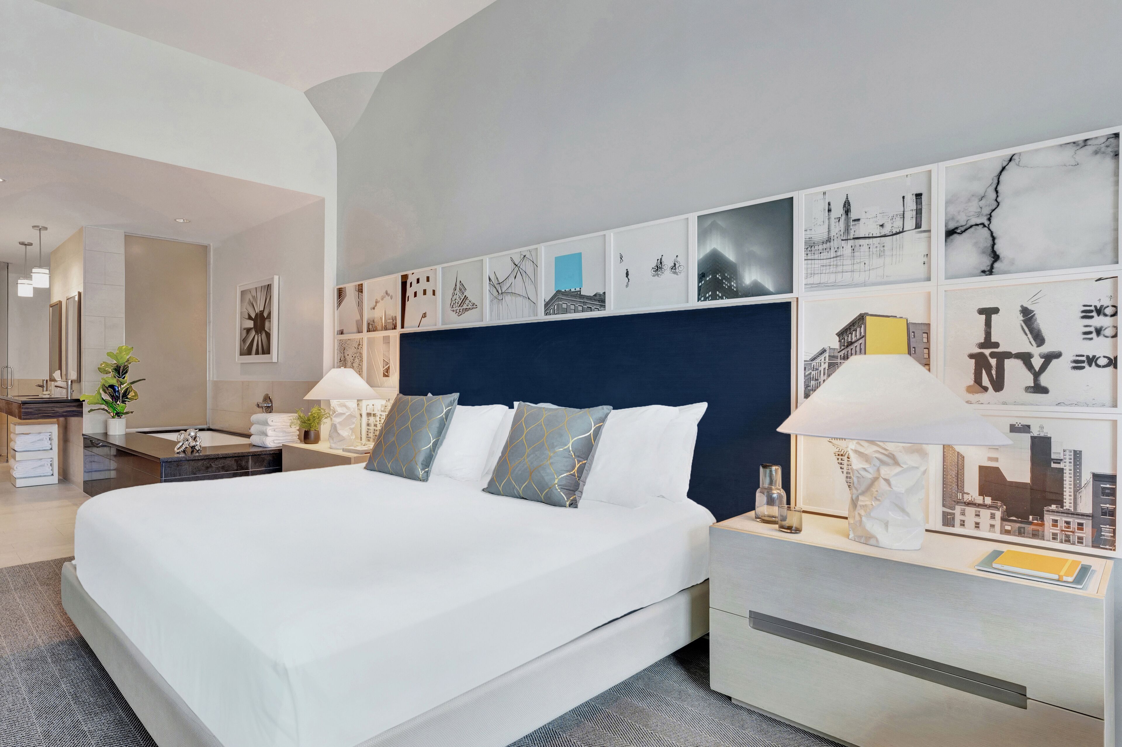 A modern hotel room with a large bed, a headboard with artwork, a bedside table with a lamp, and a nightstand with a lamp.