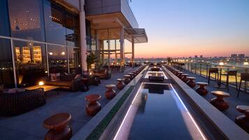 Une terrasse sur le toit avec des sièges extérieurs, un long bassin d’eau aux reflets changeants et une vue sur les toits de la ville au crépuscule.
