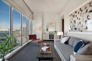 Een moderne hotelkamer met grote ramen, een bank, een bijzettafeltje met boeken, een bureau met een lamp en een kunstwerk aan de muur.