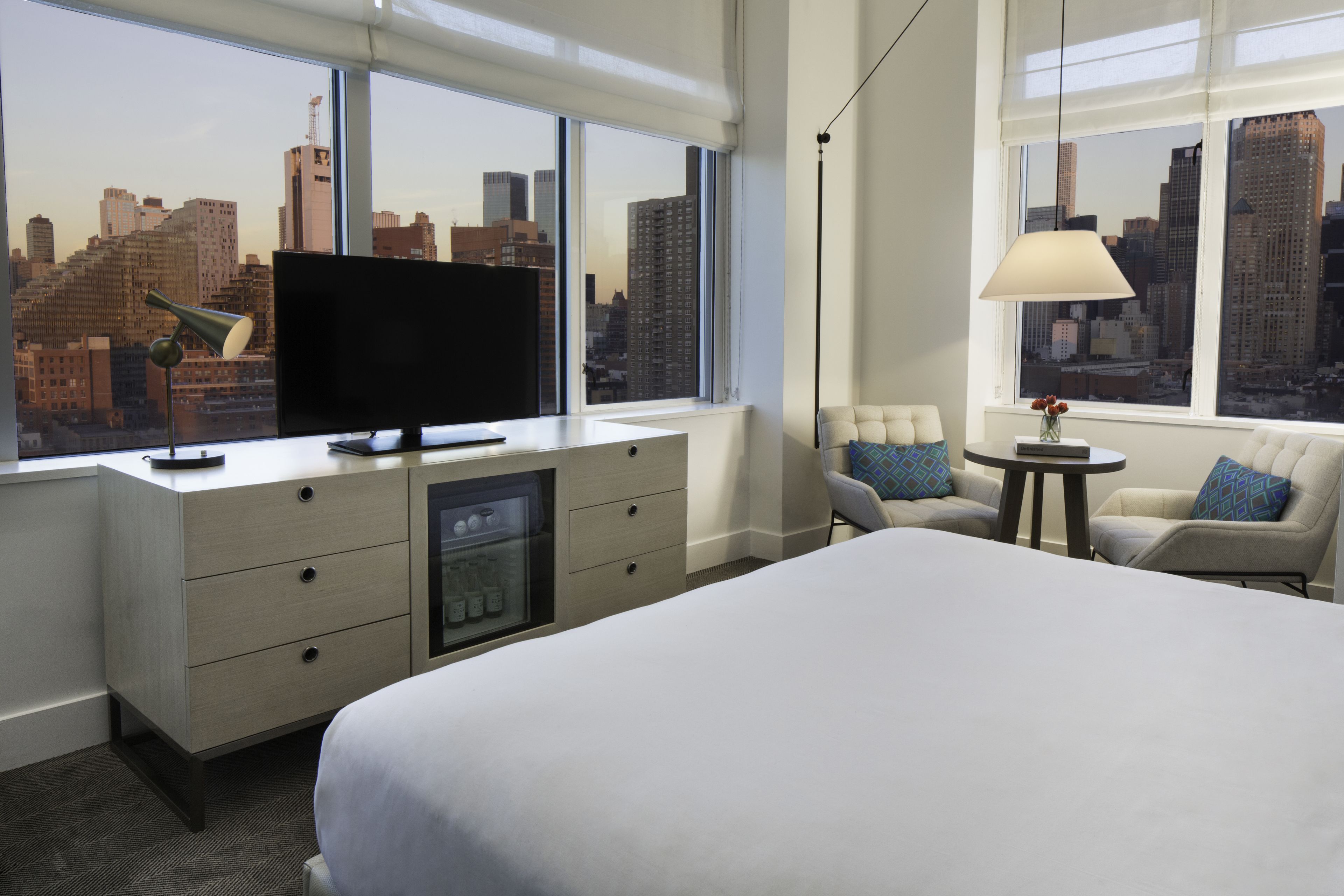 Habitación de hotel con cama, escritorio con televisor, una silla, una mesita de noche con lámpara y amplias ventanas con vista a la ciudad.