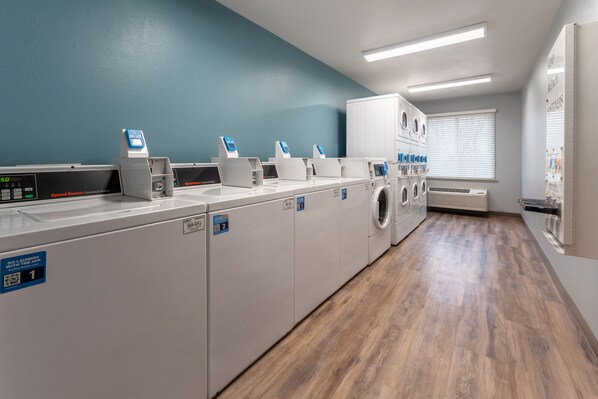A laundry facility with multiple washing machines and dryers, a digital scale, and a QR code for self-service. - WoodSpring Suites Macon West I-475 (Macon)