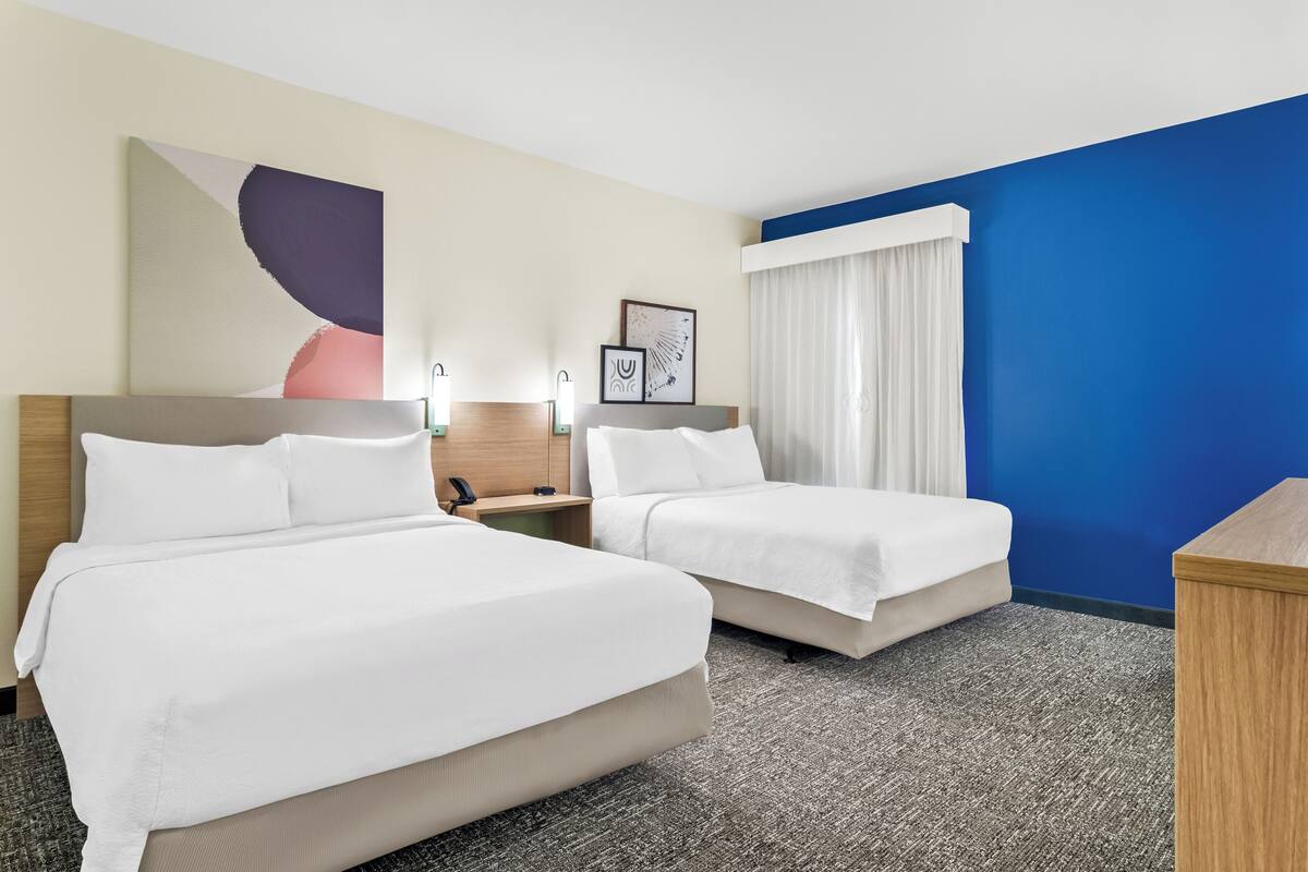 Ein Hotelzimmer mit zwei Betten, einem Schreibtisch und einem großen, abstrakten Gemälde an der Wand.