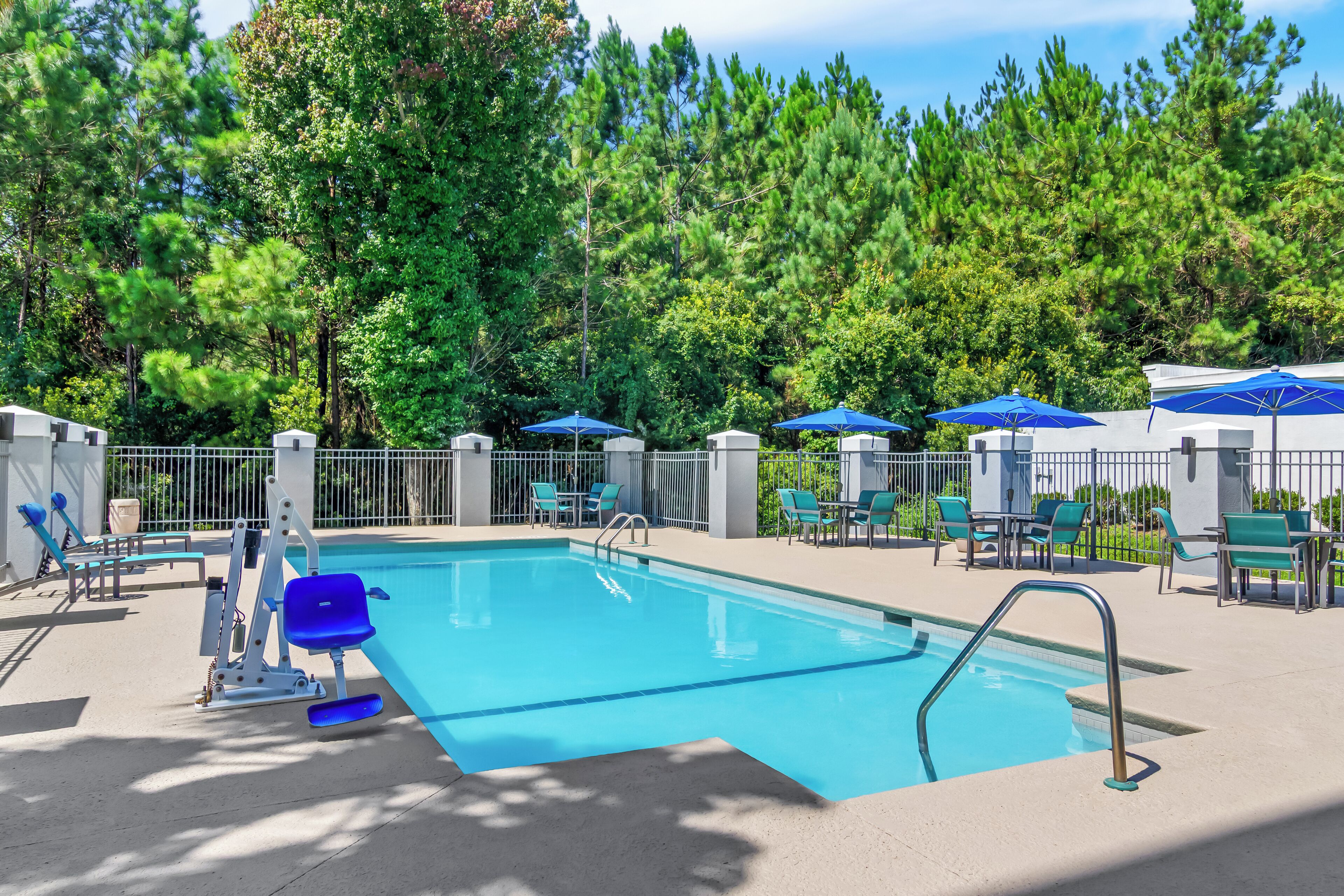Une piscine rectangulaire avec un escalier en métal, entourée d’une clôture blanche. On y trouve des chaises et des parasols bleus, et l’ensemble est à l’ombre grâce aux arbres.