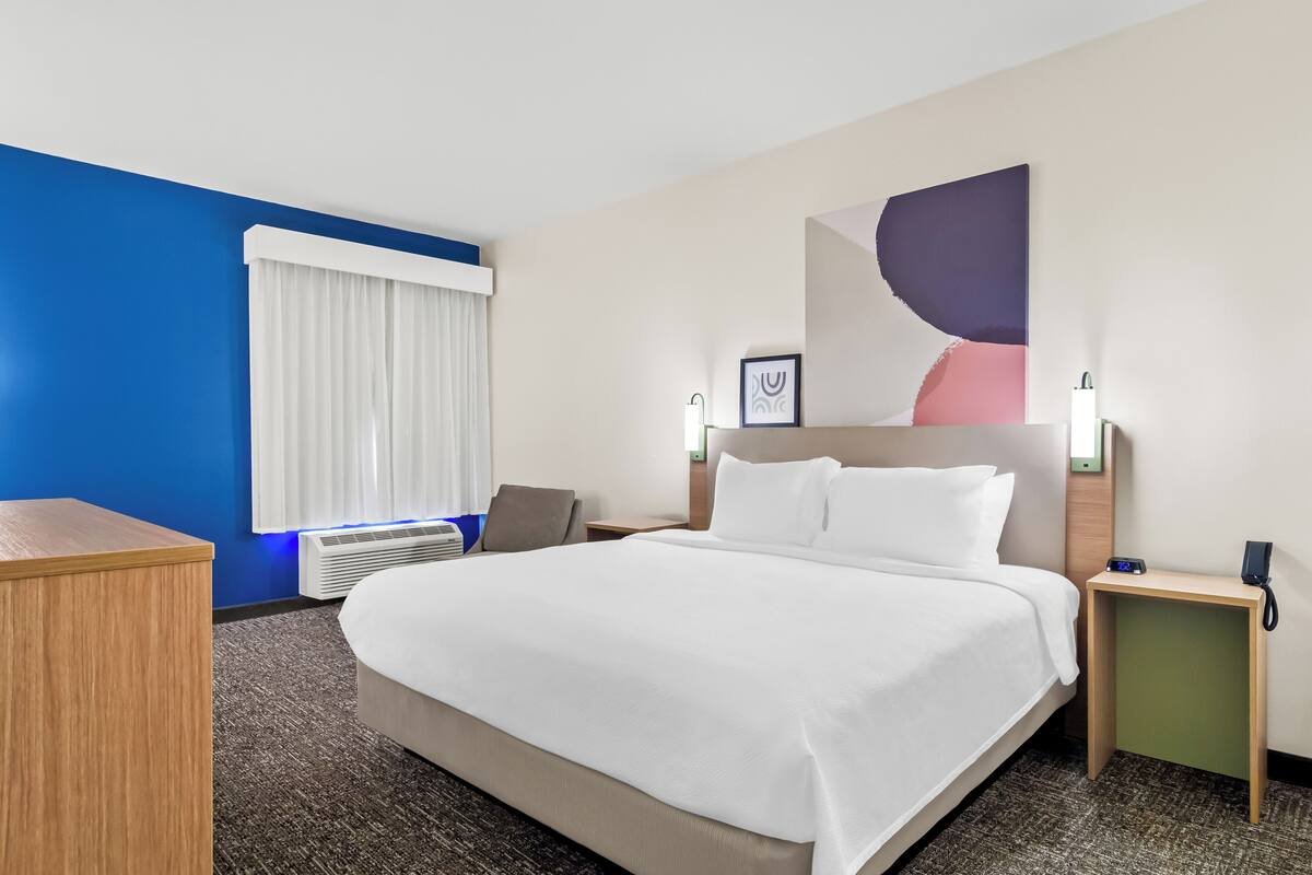 Ein Hotelzimmer mit einem großen Bett, einem Schreibtisch, einem Sessel, einem Nachttisch mit Telefon und einem an der Wand hängenden Kunstwerk.