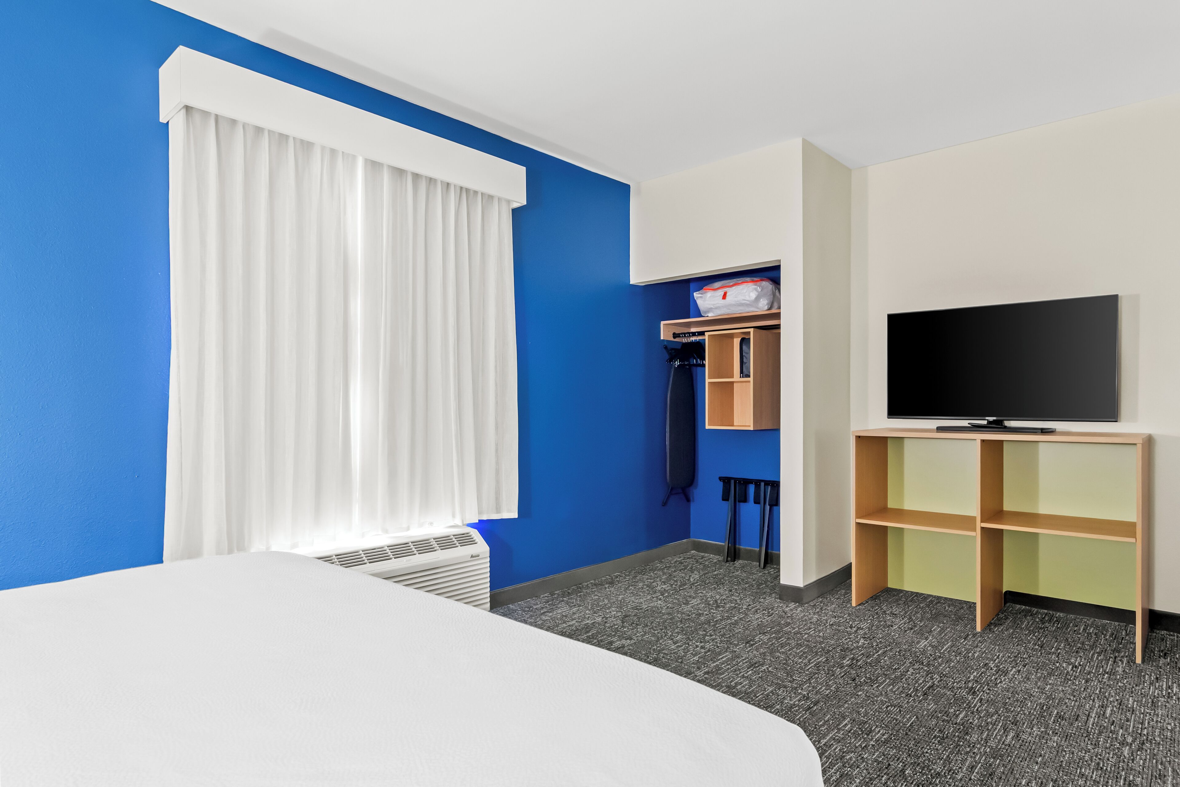 Een hotelkamer met een bed, een televisie op een standaard, een raam met witte gordijnen, een klein plankje met een tas en een verwarming.