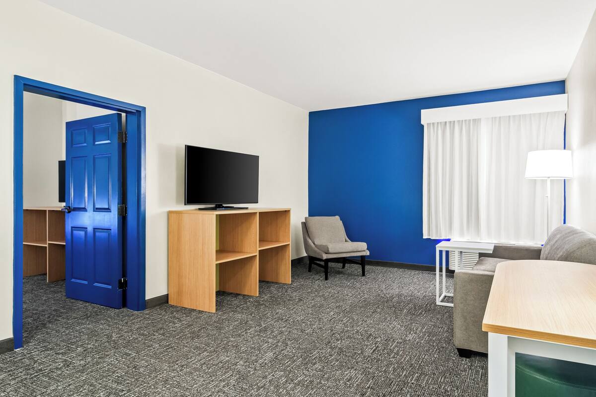 Ein Hotelzimmer mit blauer Tür, einem Fernseher auf einem Holzboden, einem grauen Sessel, einer Couch und einem Fenster mit weißen Vorhängen.
