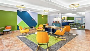 Um lobby moderno e bem iluminado, com cadeiras coloridas, uma mesa central e uma parede verde com design geométrico.