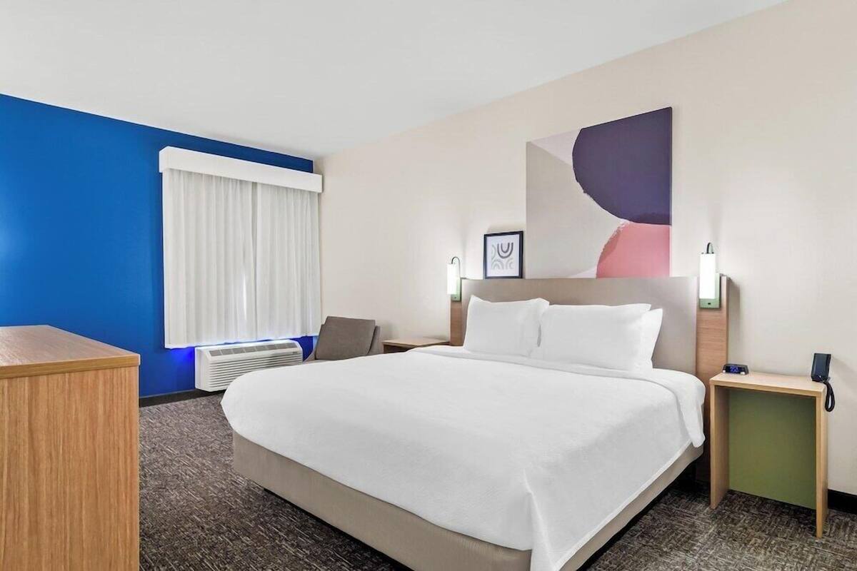 Ein Hotelzimmer mit einem Bett, einem Schreibtisch, einem Stuhl, einem Nachttisch, einem an der Wand befestigten Telefon und einem Fenster mit Vorhängen.