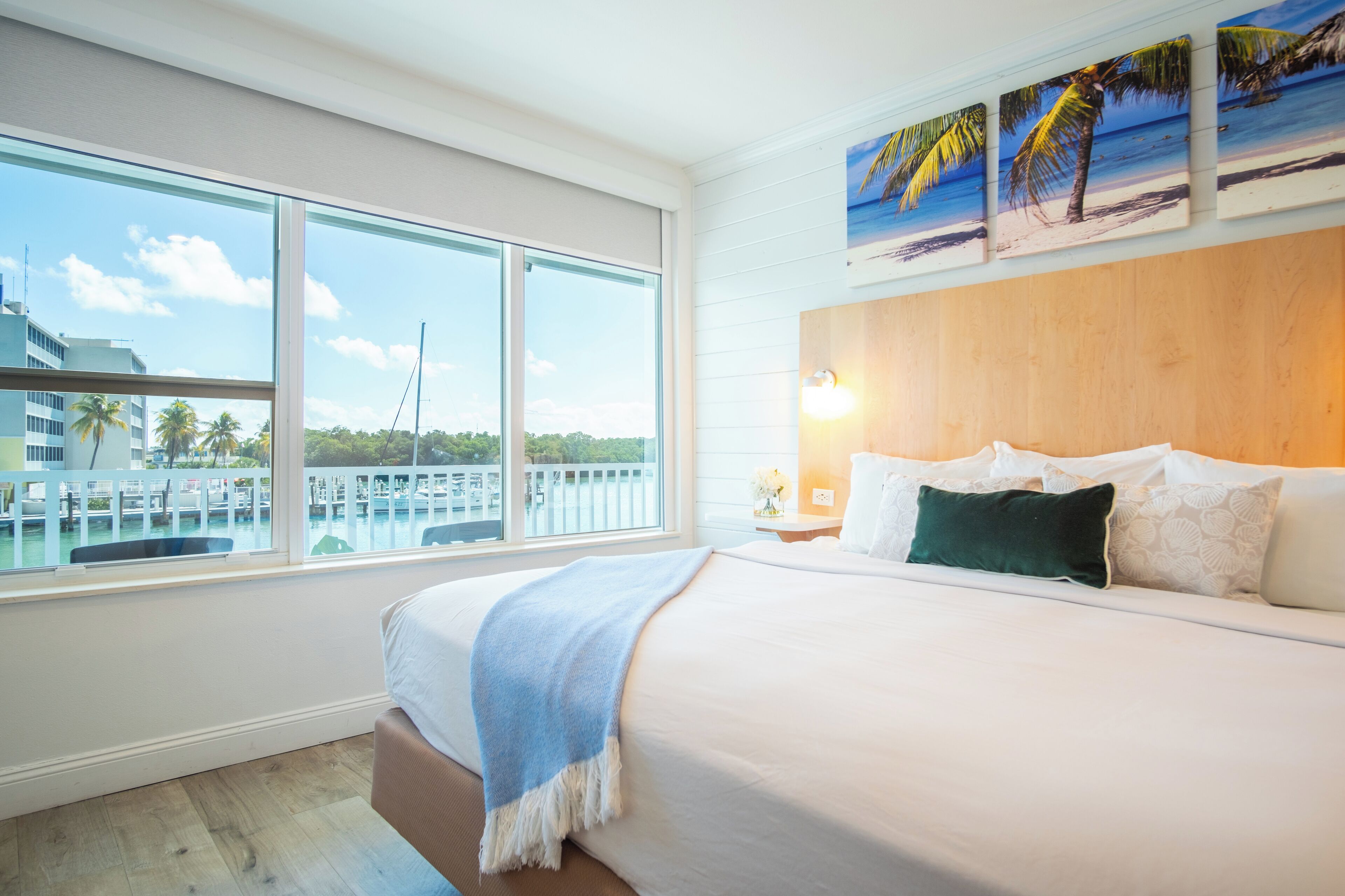 A hotel room with a large window overlooking a marina, a wooden headboard, and a bed with white linens and a blue throw blanket.