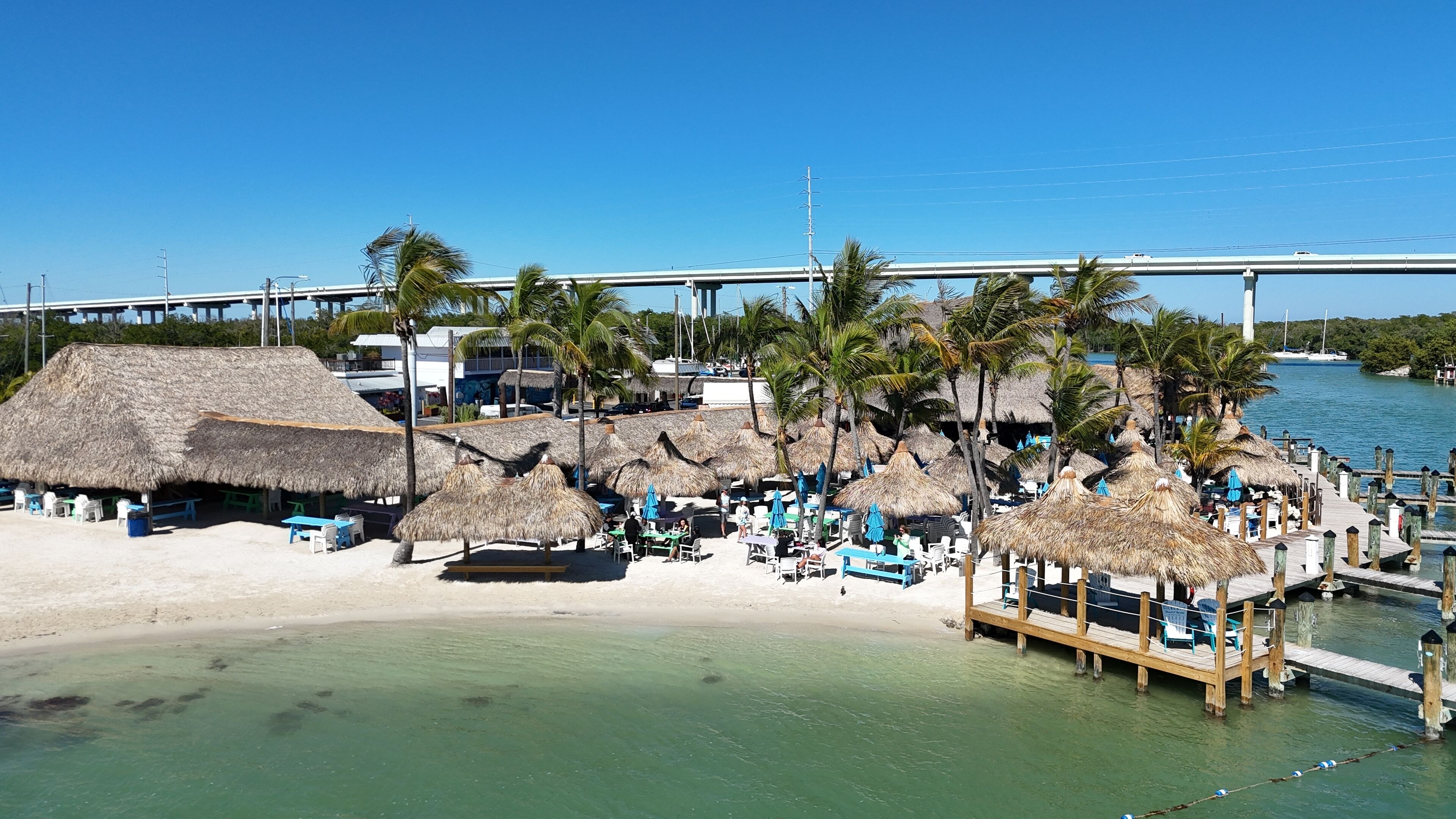 Un resort frente a la playa con cabañas de techo de paja, palmeras y un muelle que se adentra en el agua.