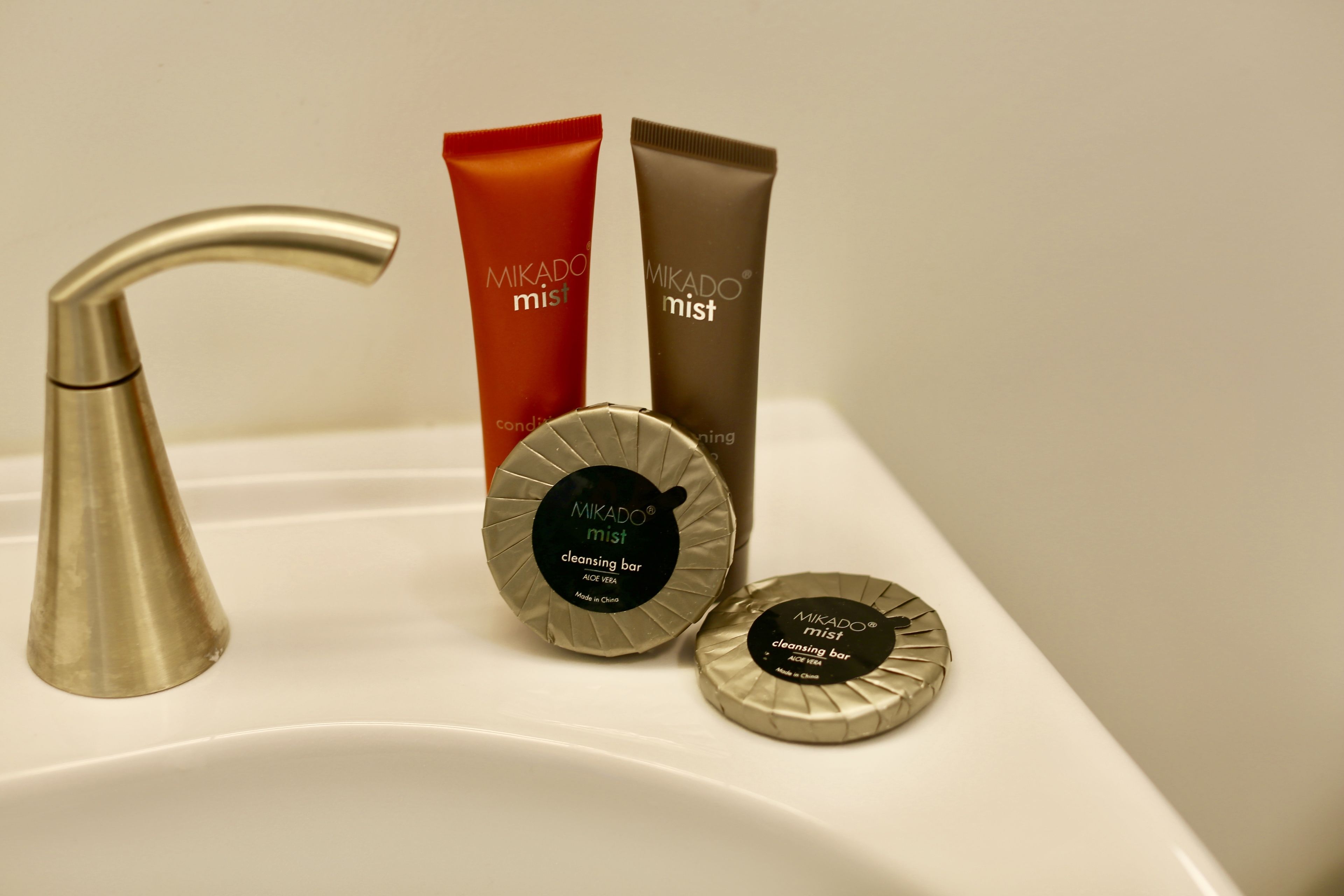 Un lavamanos de baño con un grifo moderno y tres productos Mikado mist: un tubo rojo, un tubo marrón y dos envases redondos.