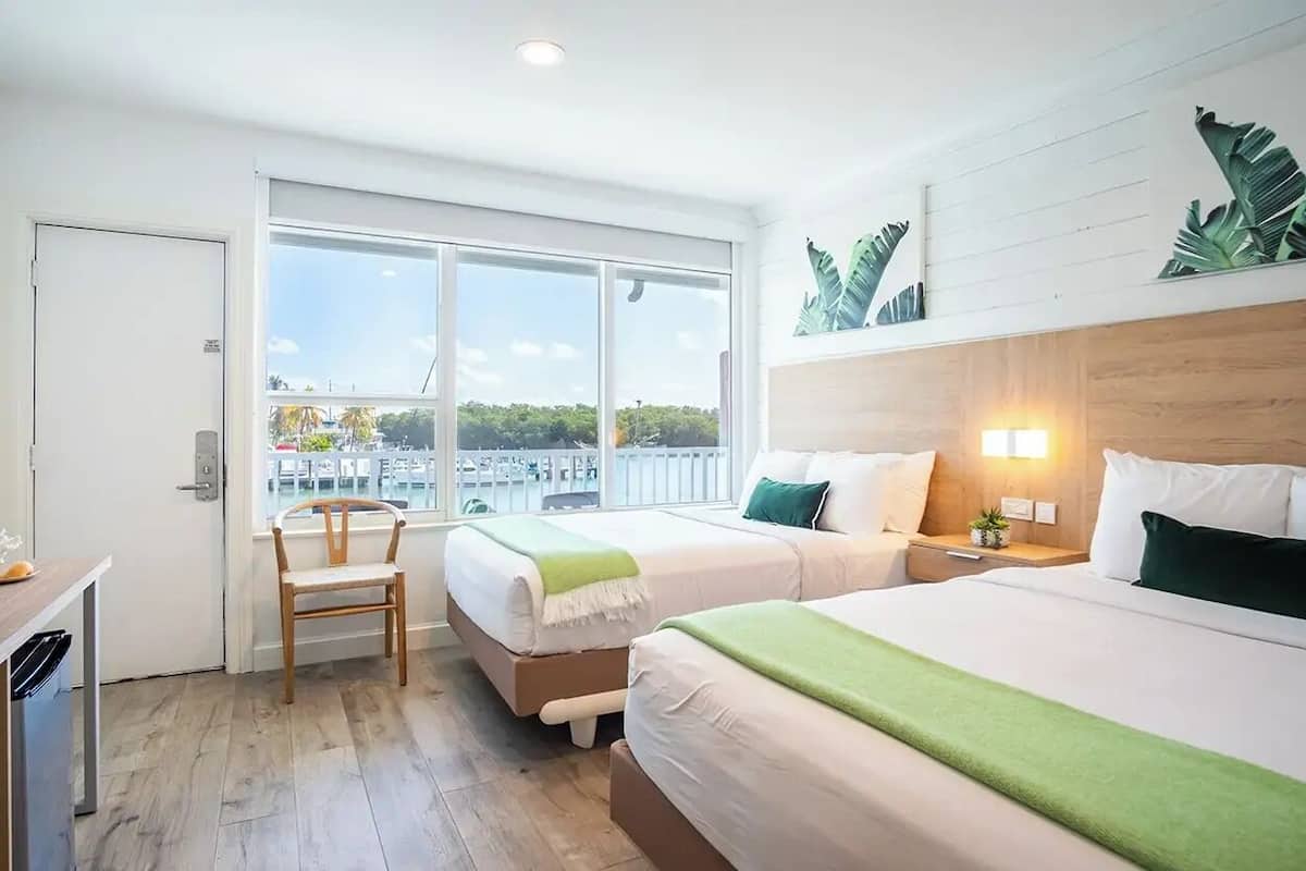 A hotel room with two beds, a wooden floor, a window with a view of the city, and a small desk with a chair.