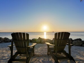 Two Adirondack chairs facing a sunset over a calm sea with rocks in the foreground.