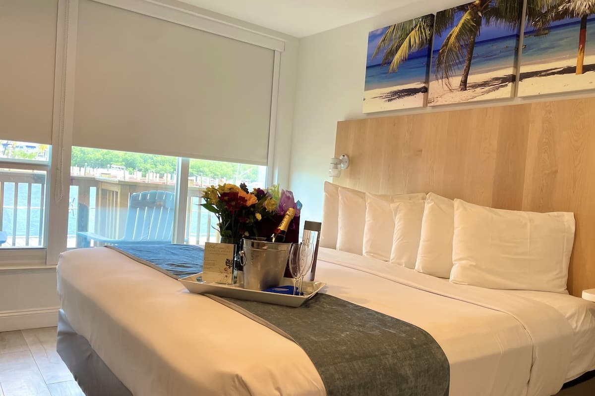 A hotel room with a bed, a bedside table with a tray of flowers, a bottle of champagne, and a painting of a beach scene.