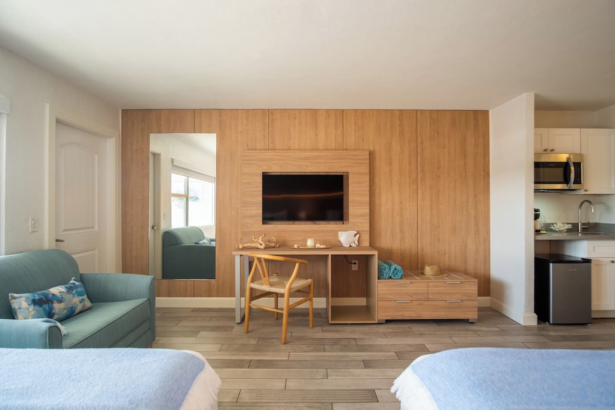 A modern hotel room with a wooden accent wall, a flat-screen TV, a desk with a chair, a bed, a sofa, and a kitchenette.