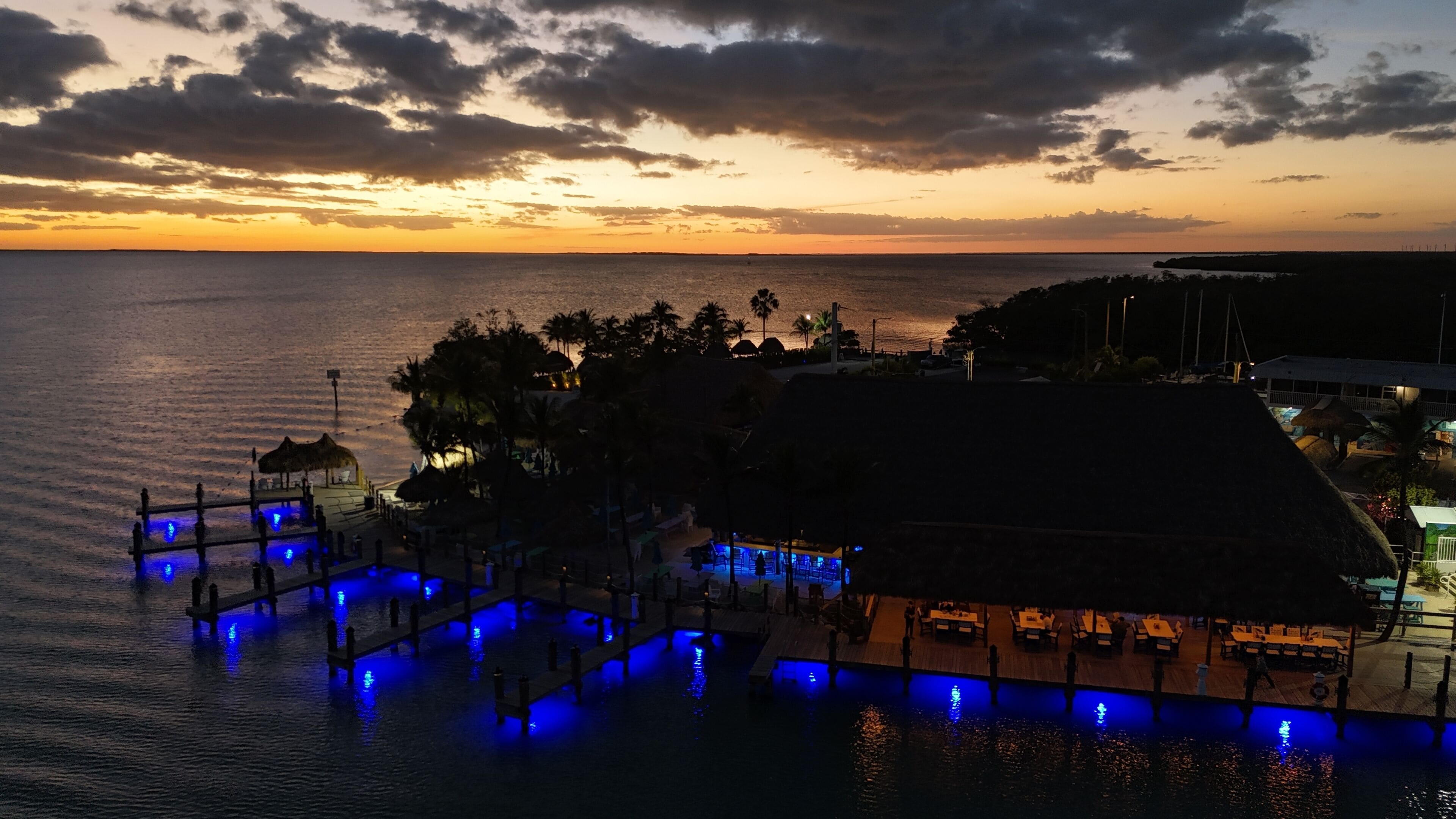 Ein Strandresort mit Poolbereich, beleuchtet durch blaues Licht, einem Essbereich und Blick auf den Sonnenuntergang über dem Meer.