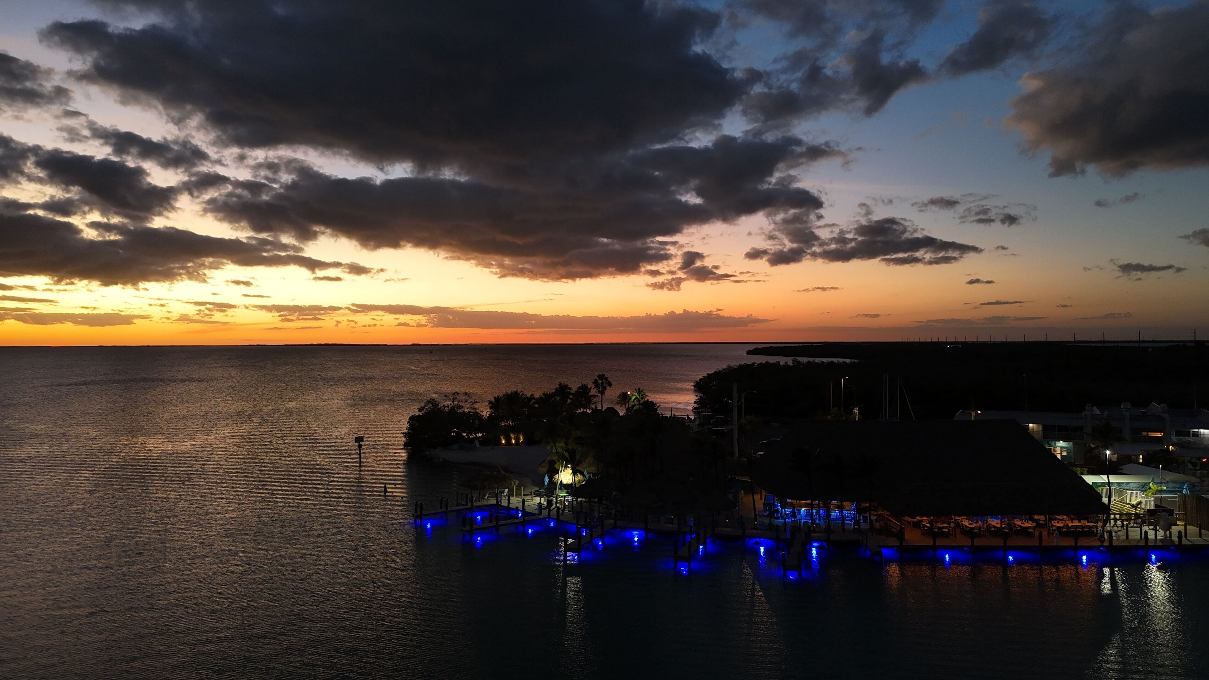 Vista al agua con un atardecer, un muelle con luces azules y una costa lejana.