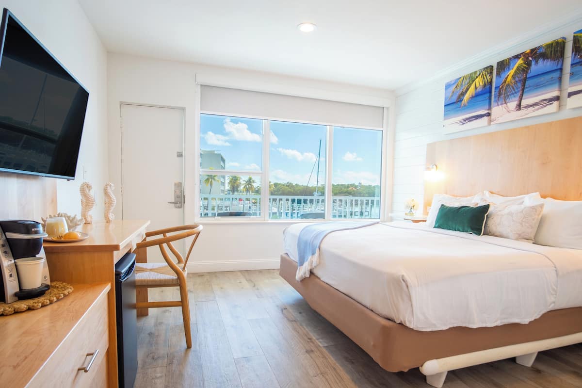 A hotel room with a bed, a TV, a desk with a chair, a window with a view of the sea, and a painting on the wall.