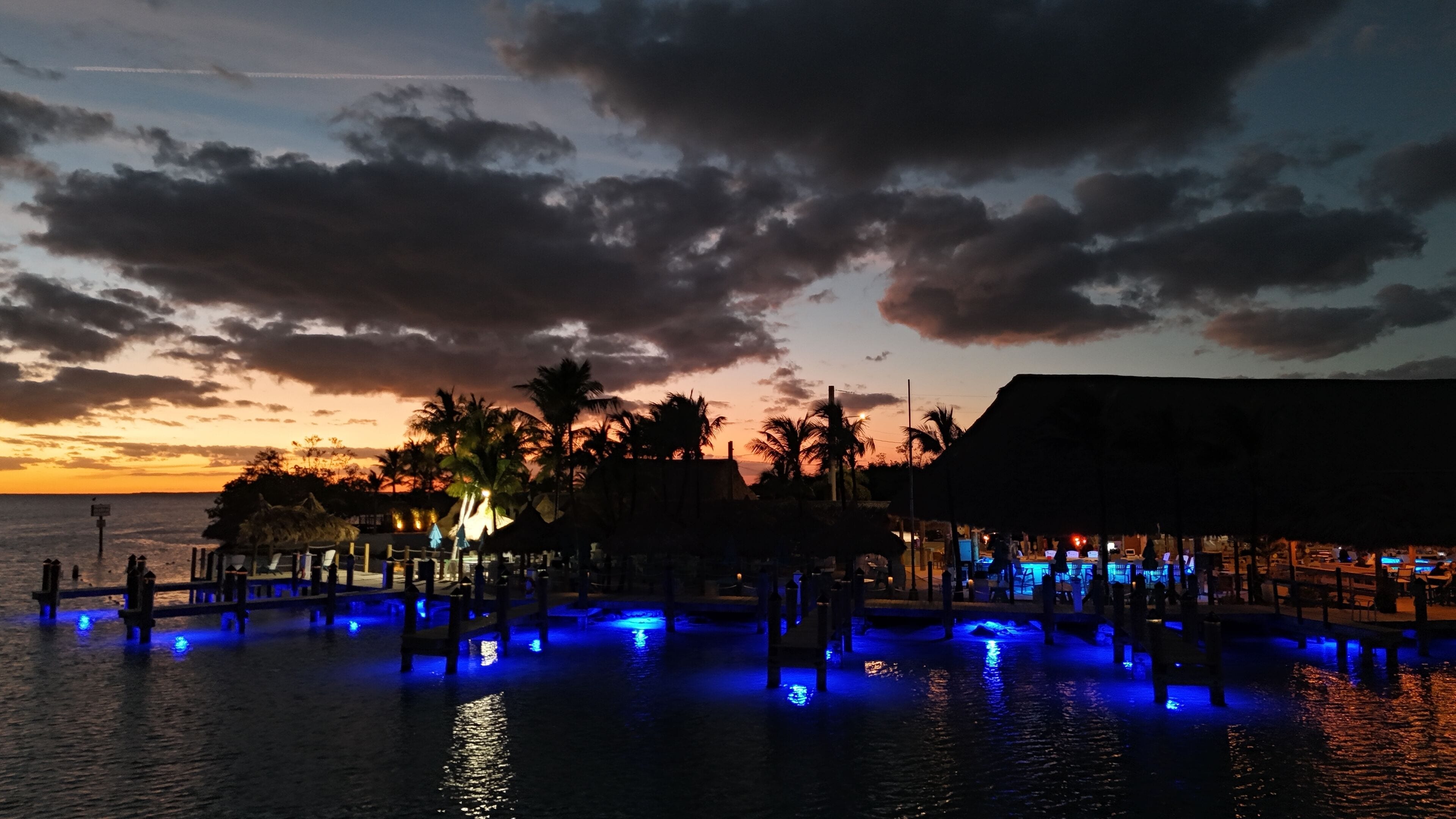 A waterfront resort with a pool area illuminated by blue lights, palm trees, and a sunset in the background.