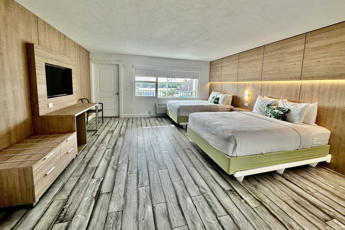 A bedroom with two beds, a TV mounted on the wall, a window with a view, and a wooden floor.