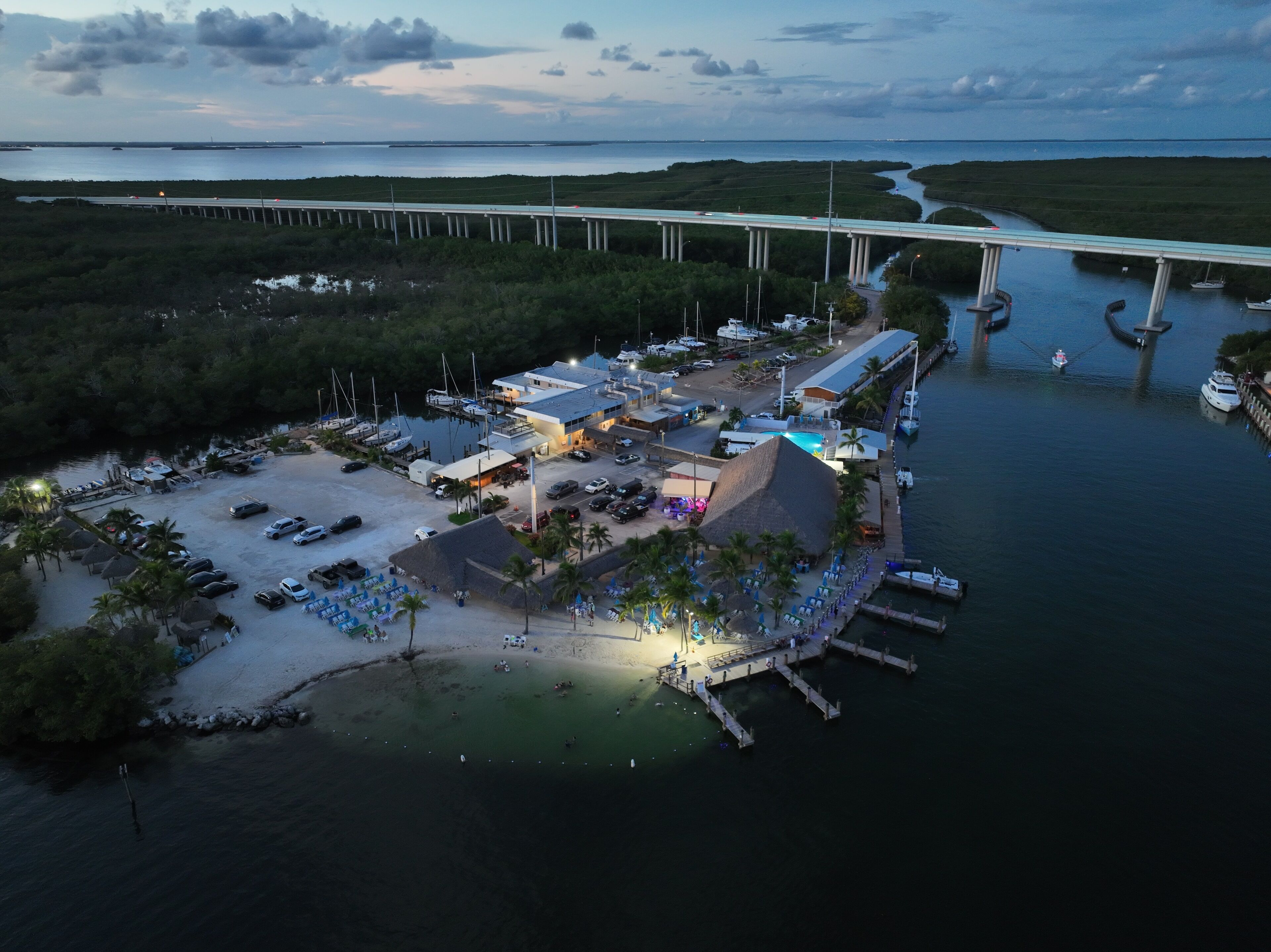 An aerial view of a coastal resort with a marina, a bridge in the background, and a large building with a pool.