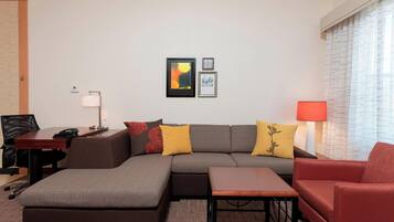 A modern living room with a sectional sofa, a red armchair, a small table, and a desk with a lamp. There are two framed artworks on the wall.