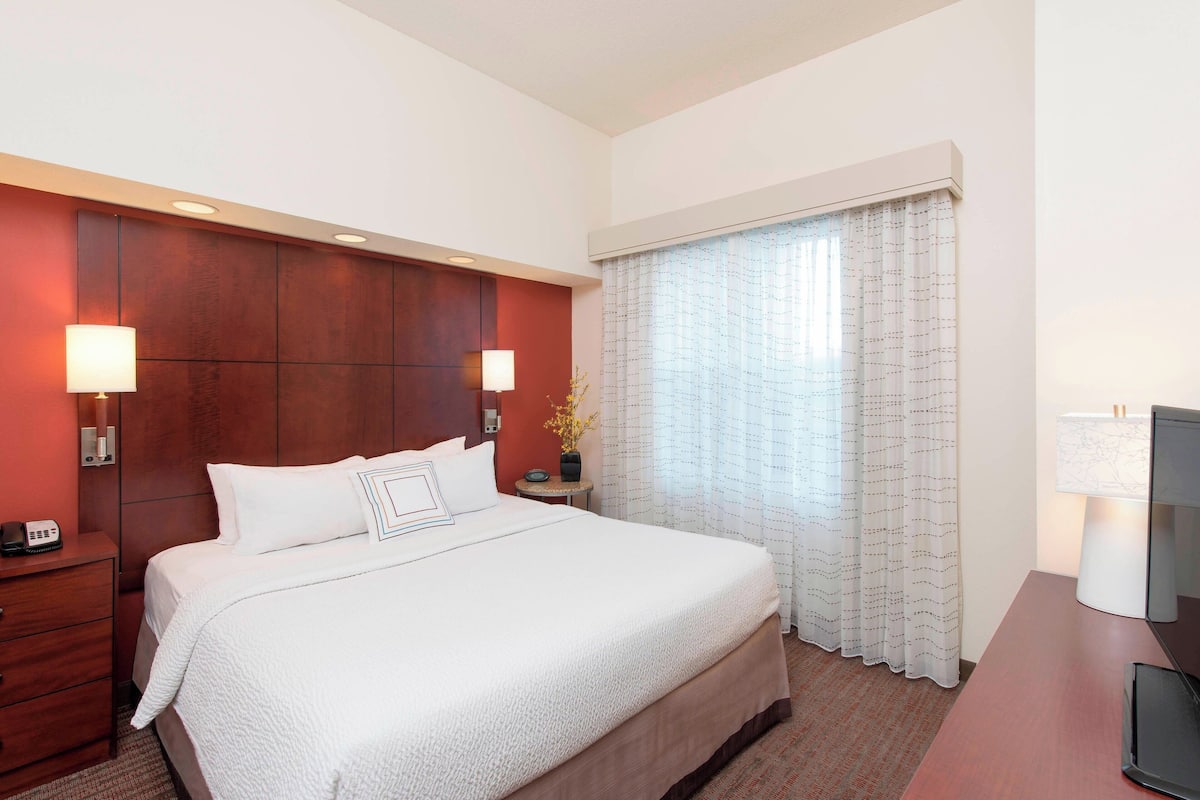 Quarto de hotel com uma cama grande, duas luminárias de cabeceira, uma escrivaninha com um computador e uma janela com cortinas.