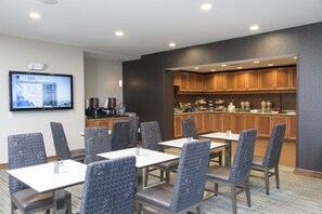 A hotel dining area with tables and chairs, a kitchen in the background, and a TV displaying weather information. - Residence Inn Marriott Moline (Moline)