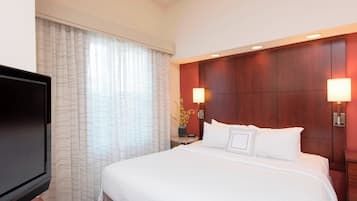 Ein Hotelzimmer mit einem großen Bett, einem Flachbildfernseher, einem Nachttisch mit Lampe und einem Fenster mit Vorhängen.
