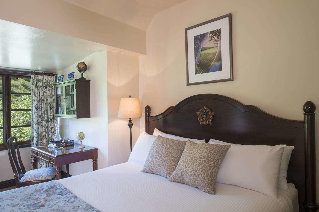 A neatly made bed with a dark headboard, a desk with a lamp, a globe, and a window with curtains.