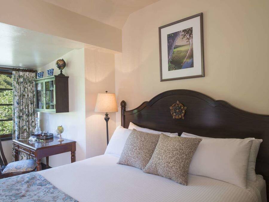 A neatly made bed with a dark headboard, a desk with a lamp, a globe, and a window with curtains.