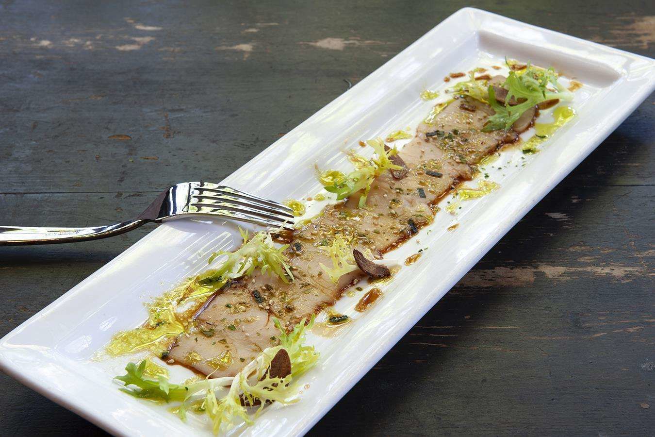 Un plato rectangular blanco con un bocado de comida, adornado con hojas verdes y un hilo de salsa, sobre una superficie de madera oscura.