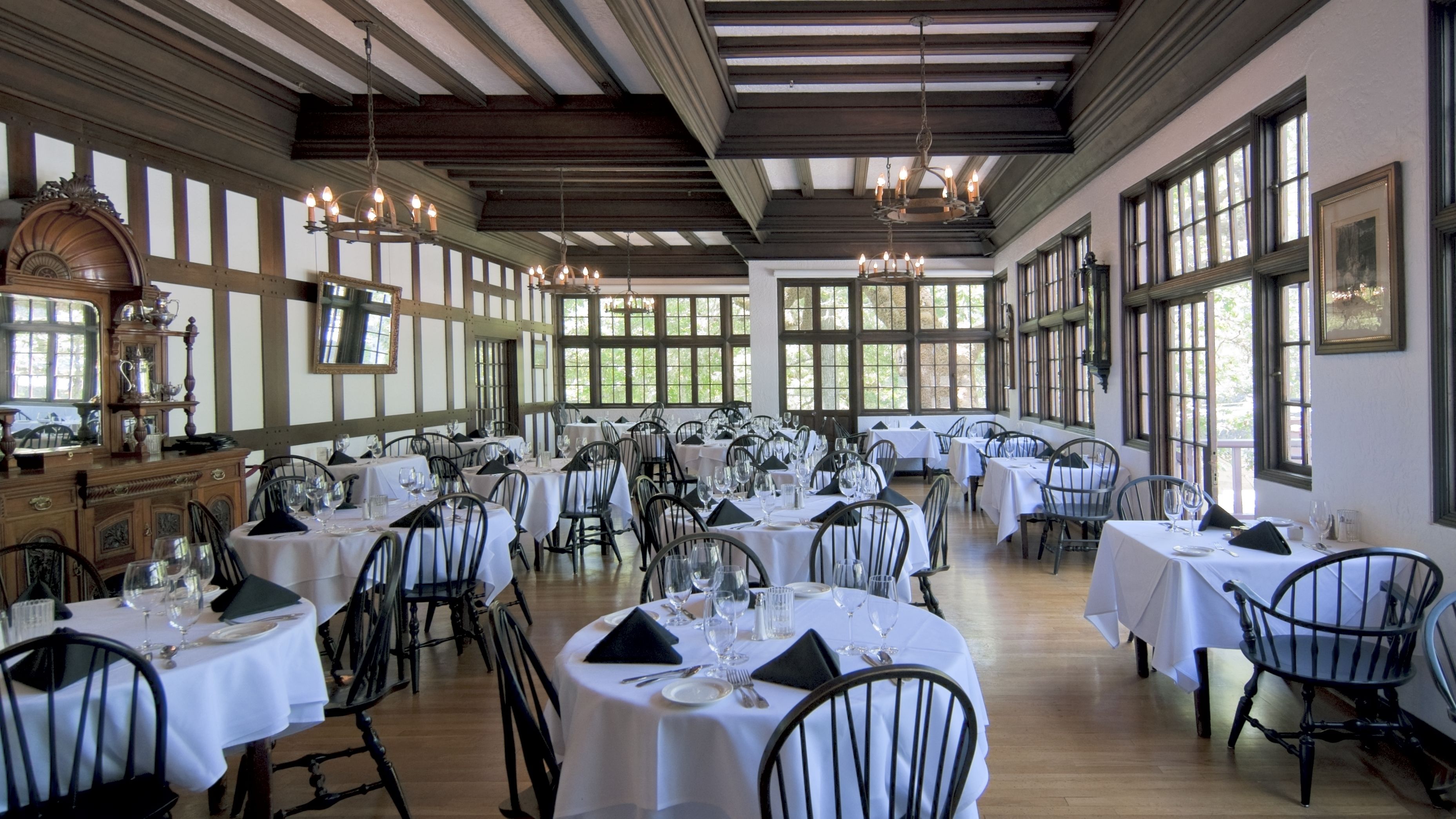 Une vaste salle à manger avec de nombreuses tables dressées pour le repas, agrémentée de poutres en bois au plafond et de grandes fenêtres.