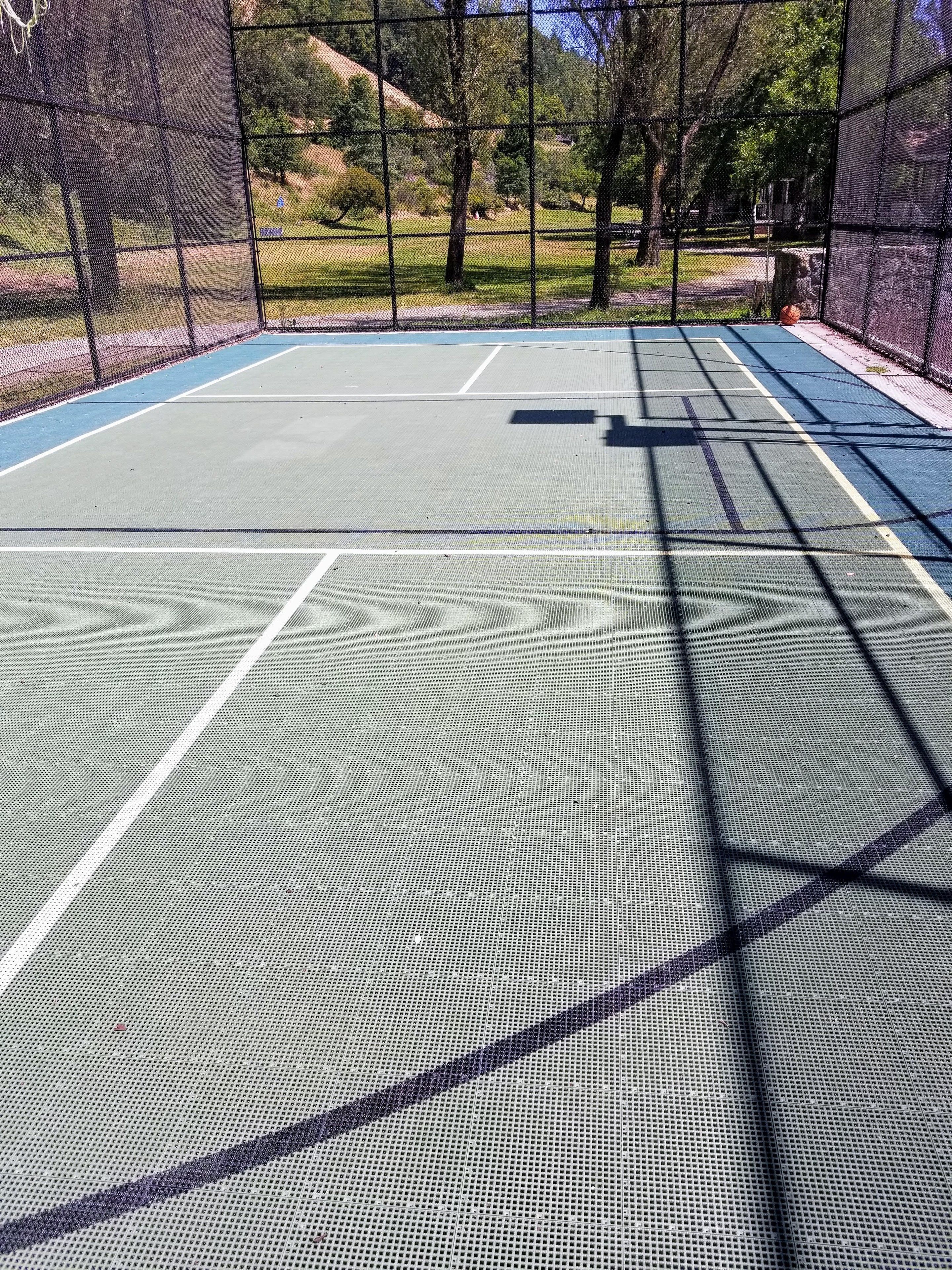 An outdoor tennis court with a net, surrounded by trees and a fence.