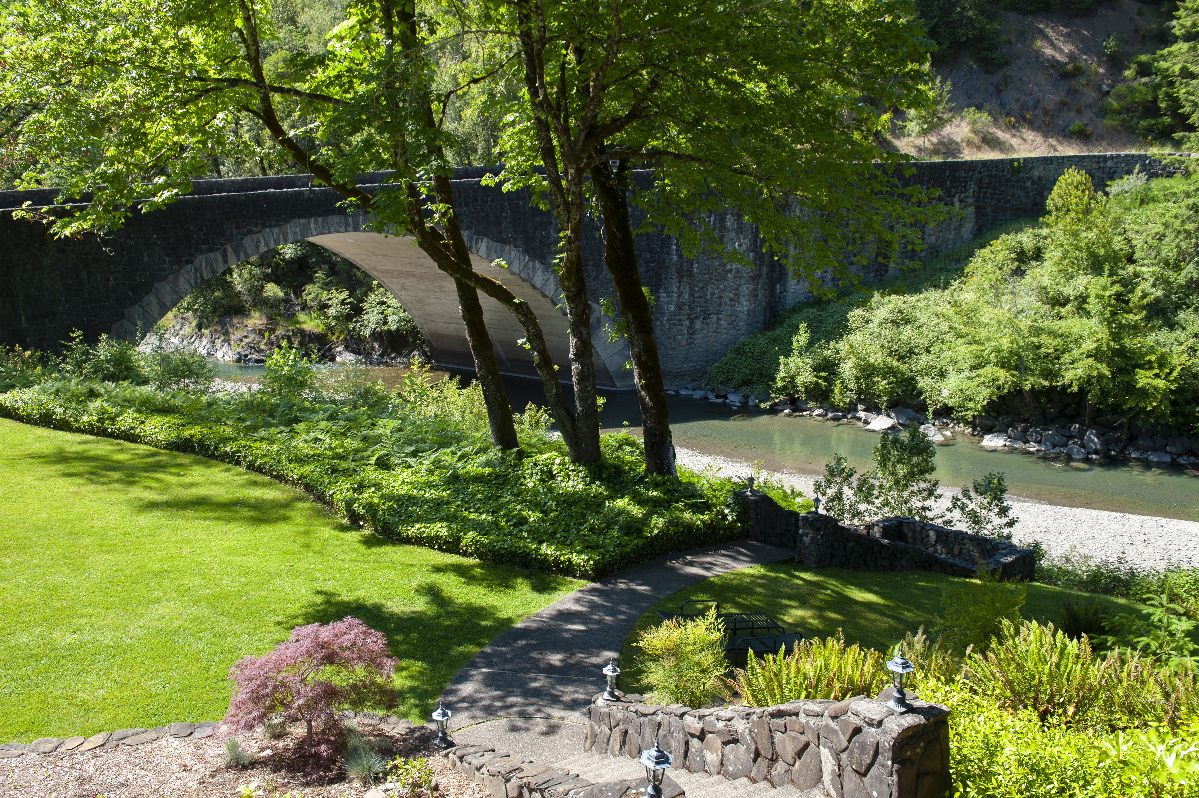 A stone bridge over a river, surrounded by lush greenery and a well-maintained lawn.