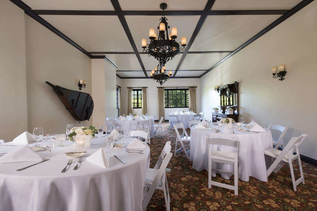 A banquet hall with round tables set for dining, a chandelier, and a piano against the wall.