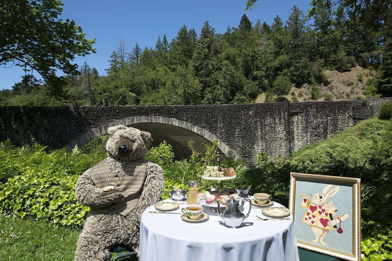 Un puente de piedra, un osito de peluche y una mesa servida con té y pasteles en un jardín.