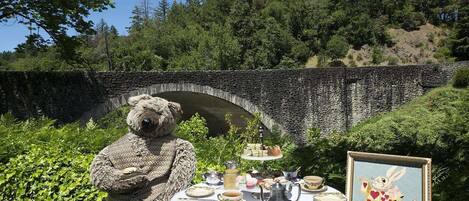 Un puente de piedra, un osito de peluche y una mesa servida con té y pasteles en un jardín.