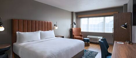 A hotel room with a large bed, a desk with a chair, a chair, a lamp, and a window with a view.