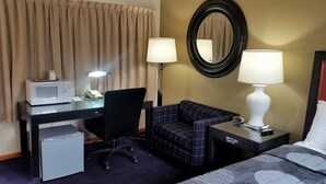 Ett hotellrum med ett skrivbord, en stol, en säng, ett litet kylskåp, en mikrovågsugn, en lampa och en spegel.
