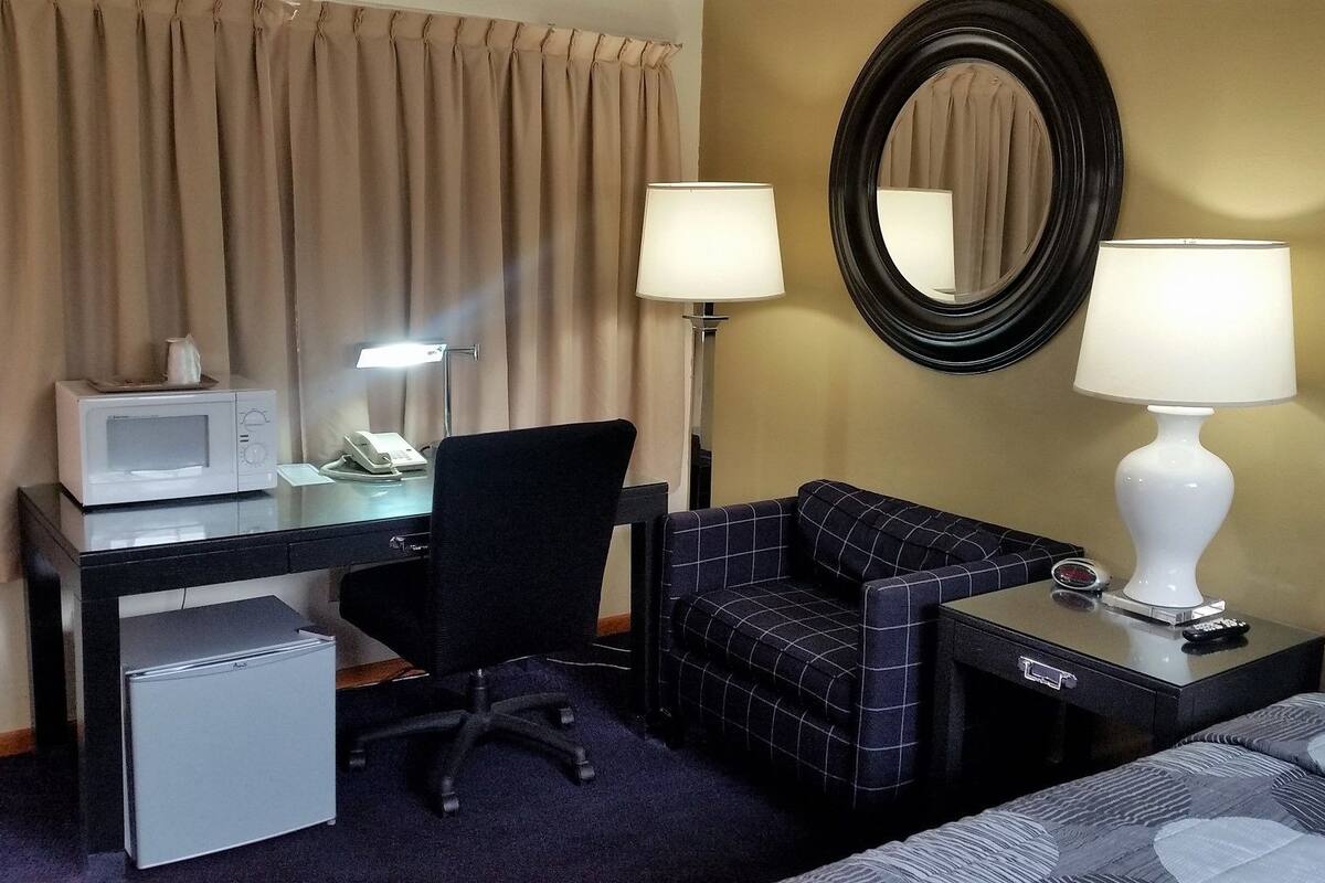 A hotel room with a desk, a chair, a bed, a small refrigerator, a microwave, a lamp, and a mirror.