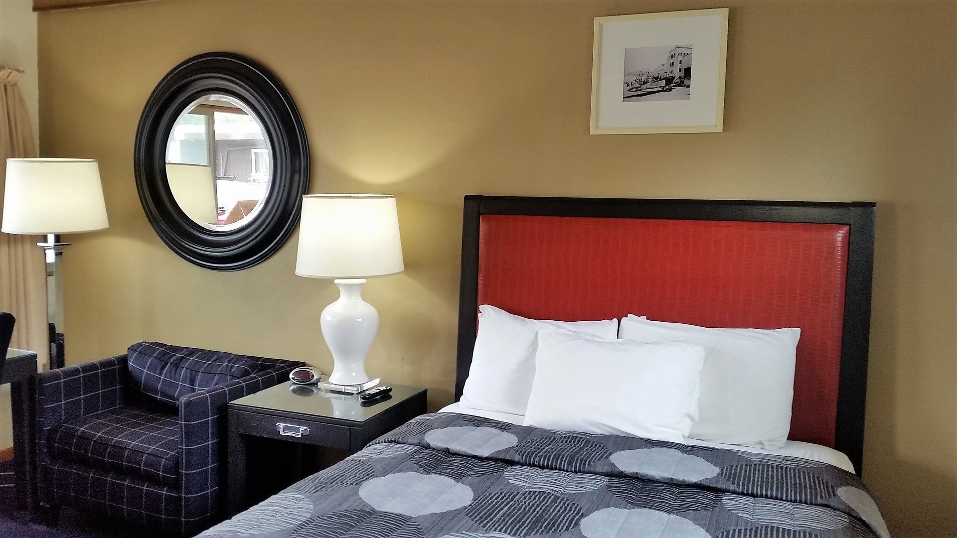 A hotel room with a bed, a nightstand, a chair, a lamp, a mirror, and a framed picture on the wall.