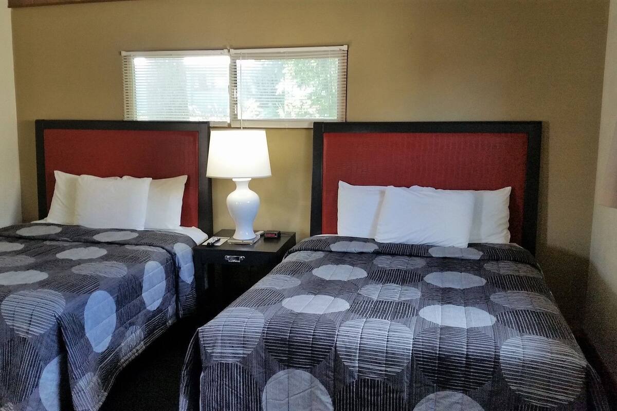 A hotel room with two beds, a nightstand with a lamp, and a window with blinds.