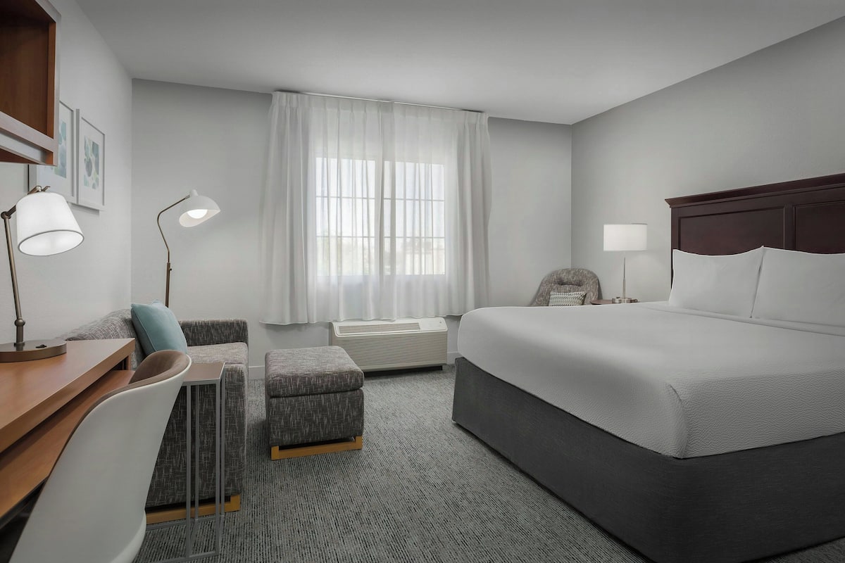 A hotel room with a bed, a desk, a chair, a sofa, a window with curtains, and a bedside table with a lamp.